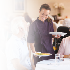 Senior living residents enjoying a meal while interacting with a staff member in a community dining room